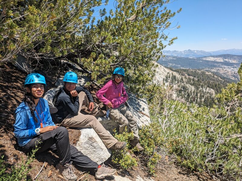 Three people wearing helmets are sitting on rocks near a cliff overlooking a valley. The people appear to be resting after some sort of activity, possibly hiking or climbing, given the helmets. The background shows a vast landscape with trees and mountains under a clear sky. The overall scene suggests an outdoor adventure in a mountainous region.